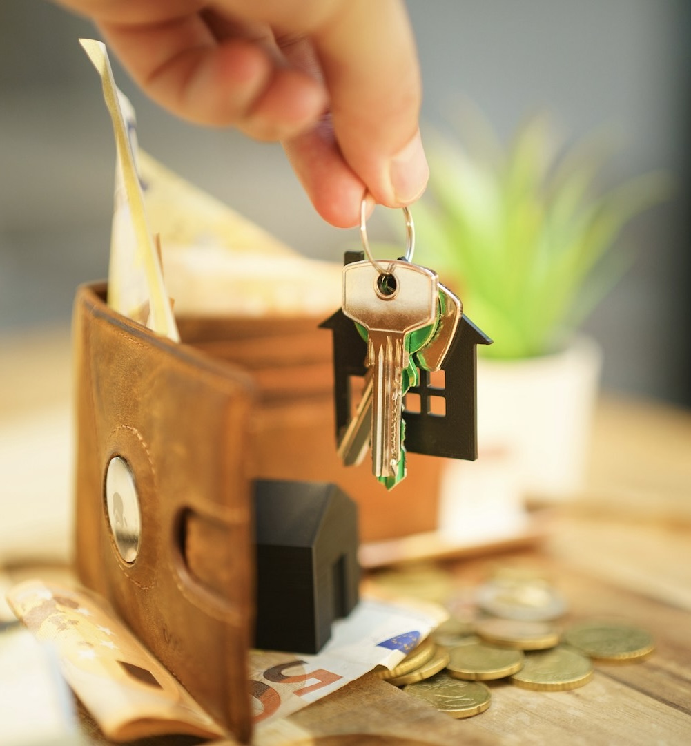 A hand holding a set of house keys above an open wallet containing euro banknotes and coins, symbolizing housing costs or property expenses.