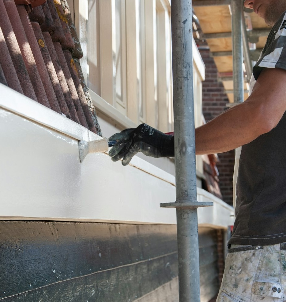 A painter is painting the exterior wooden board of a house while standing on scaffolding.